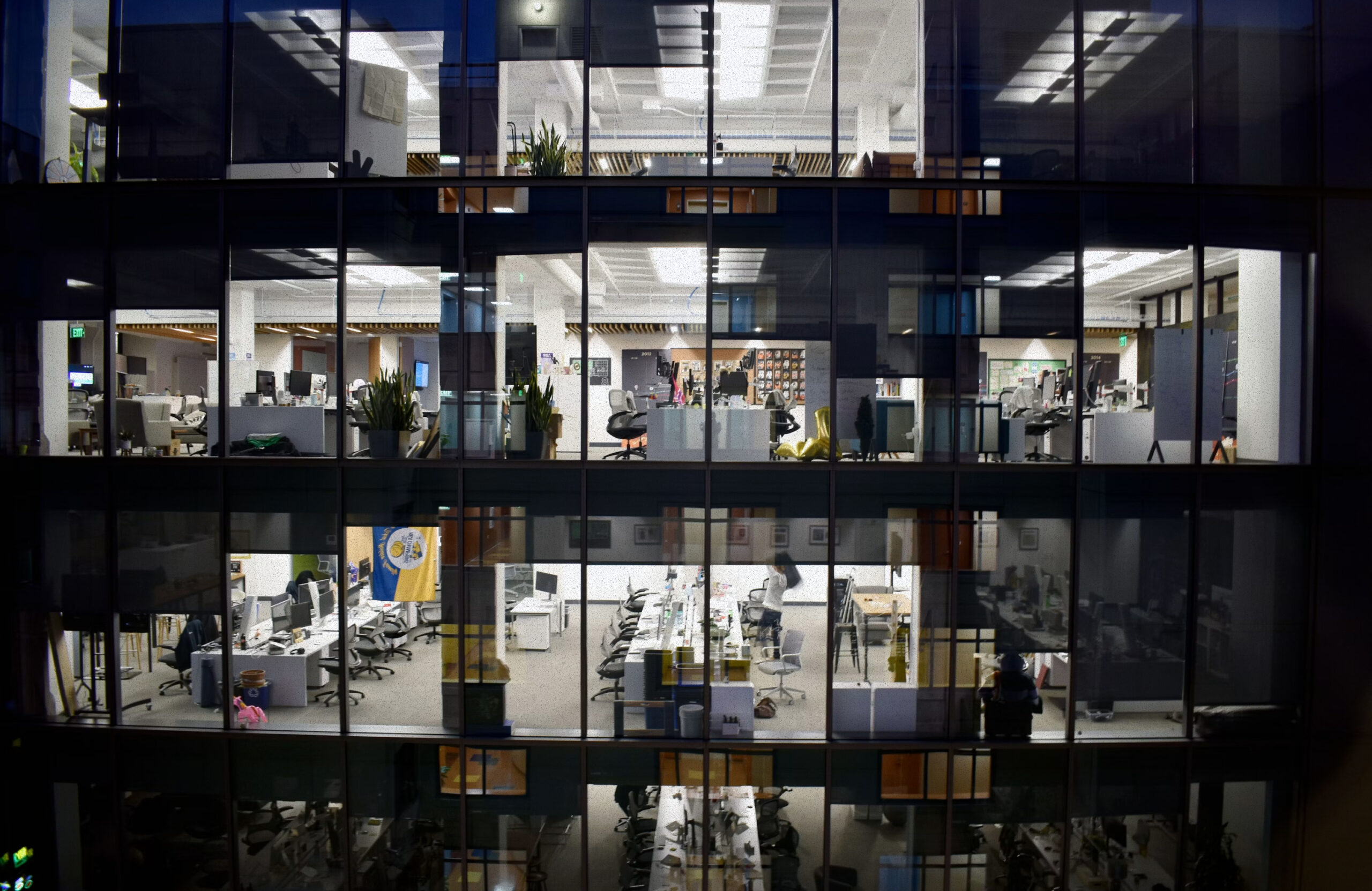 Office building with blinds open showing empty chairs and workstations.